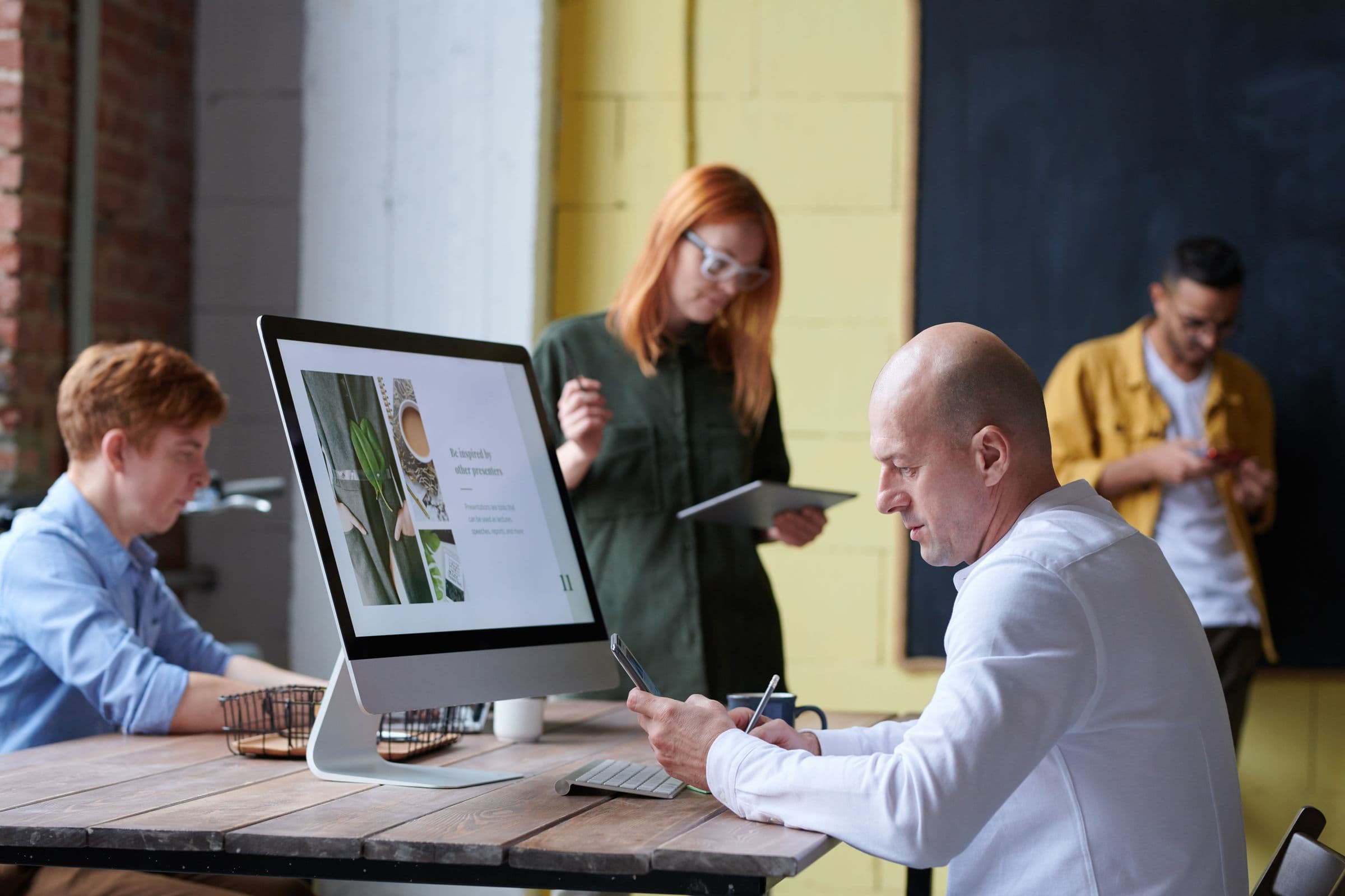People working at a shared table in a bright open-plan workspace, a large monitor showing a presentation